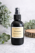 Black bottle of Xaloca Eucalyptus Room & Linen Spray with eucalyptus leaves in the background.