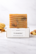 Bar of turmeric soap on a white box with 'Naloca' branding, surrounded by turmeric roots on a light background.