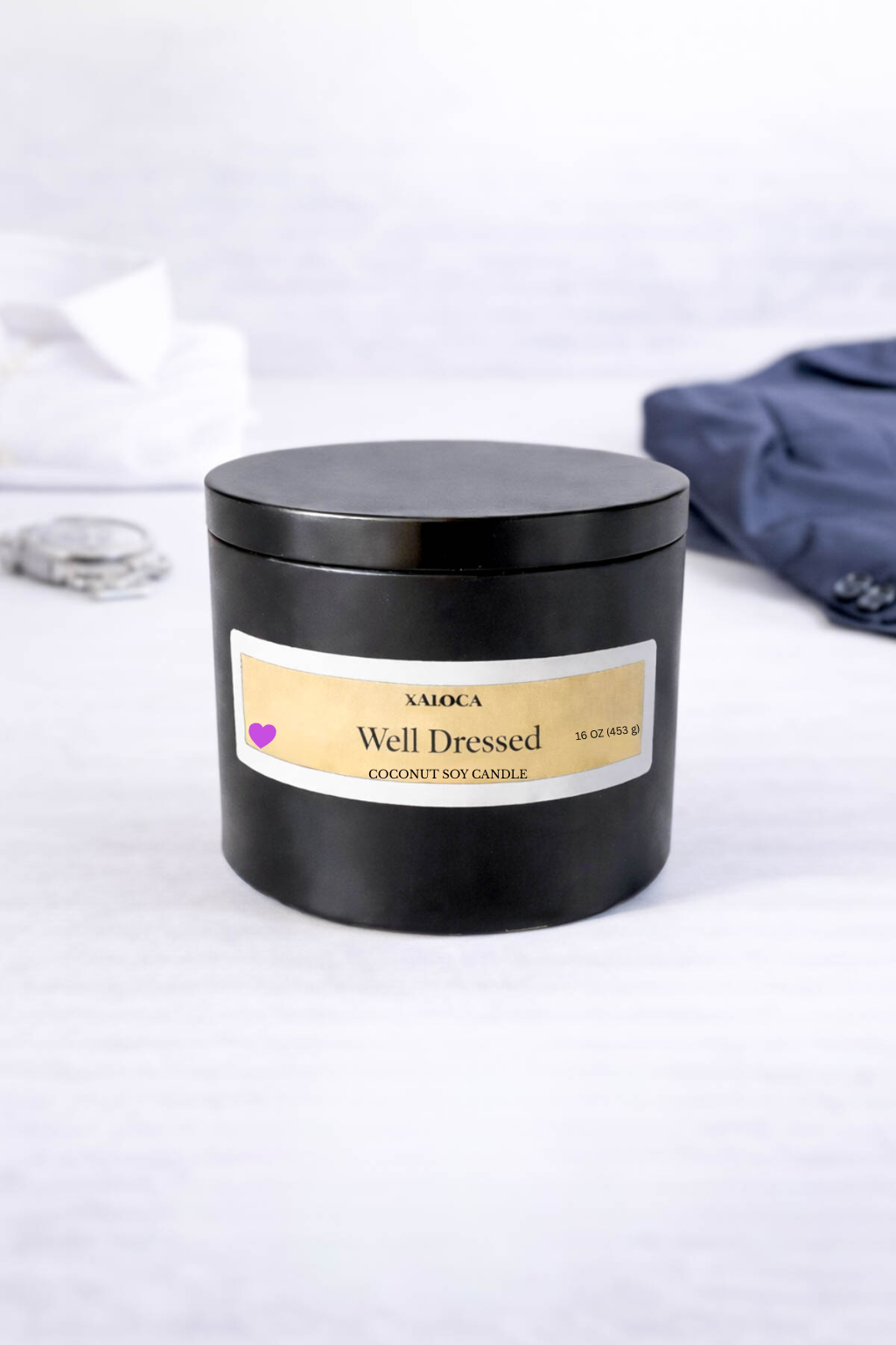 Black candle jar labeled 'Well Dressed' on a white surface with a blue garment in the background.